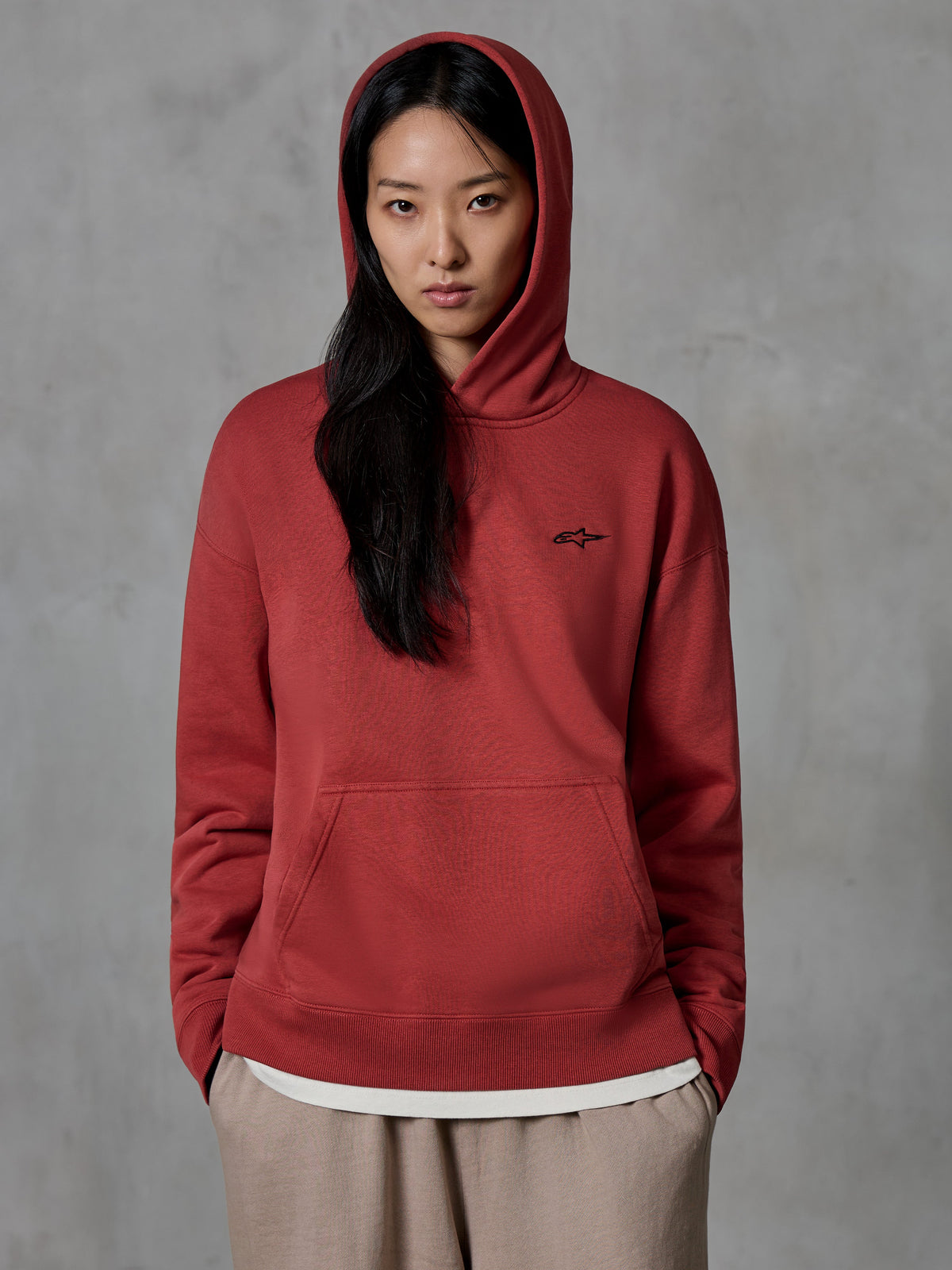 Alpinestars Essentials Ageless Hoodie, Brick red pullover sweatshirt, worn by a female model with the hood up, featuring a small black embroidered star logo on the chest, kangaroo pocket, casual motorsport-inspired lifestyle streetwear.