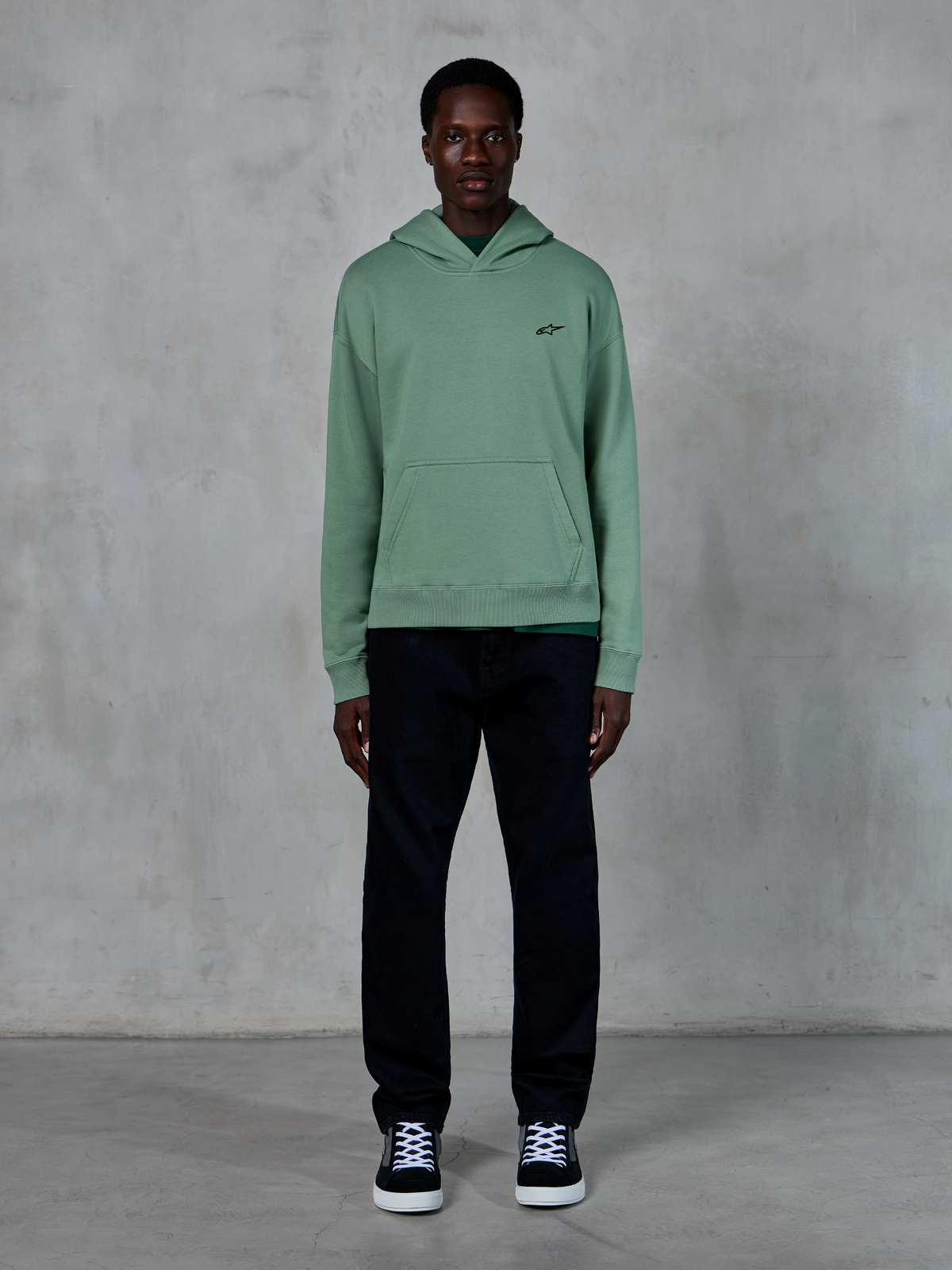 Alpinestars Essentials Ageless Hoodie, Cactus green, small black chest logo, kangaroo pocket, casual lifestyle streetwear worn with black pants and black and white sneakers against a textured wall