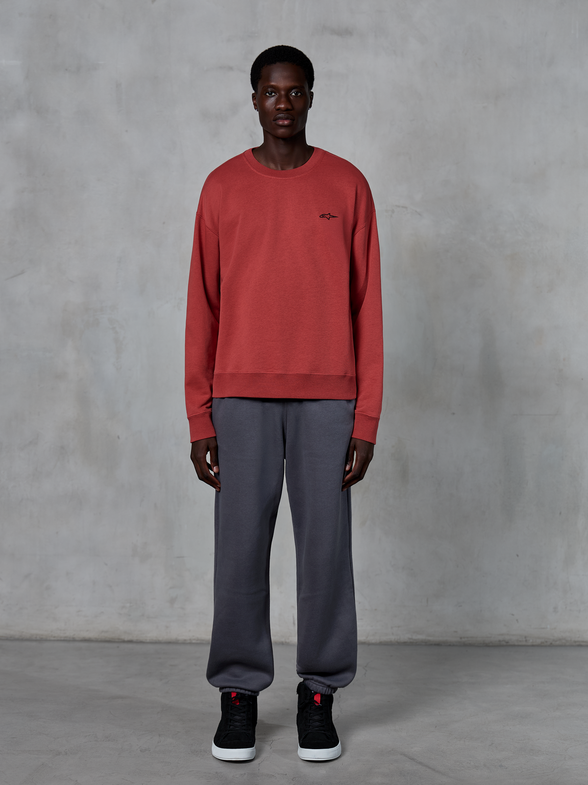 Alpinestars Essentials Ageless Crew, Brick Red crewneck sweatshirt, worn with grey sweatpants and black sneakers, featuring a small black logo on the chest, casual motorsport-inspired streetwear