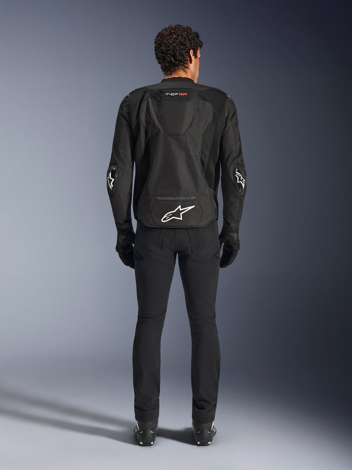 Alpinestars T-GP Air Jacket, Black, Rear view of a man wearing the textile motorcycle jacket, featuring mesh ventilation panels, an aerodynamic hump, and white Alpinestars logos on the lower back and elbows, worn with black riding pants and gloves