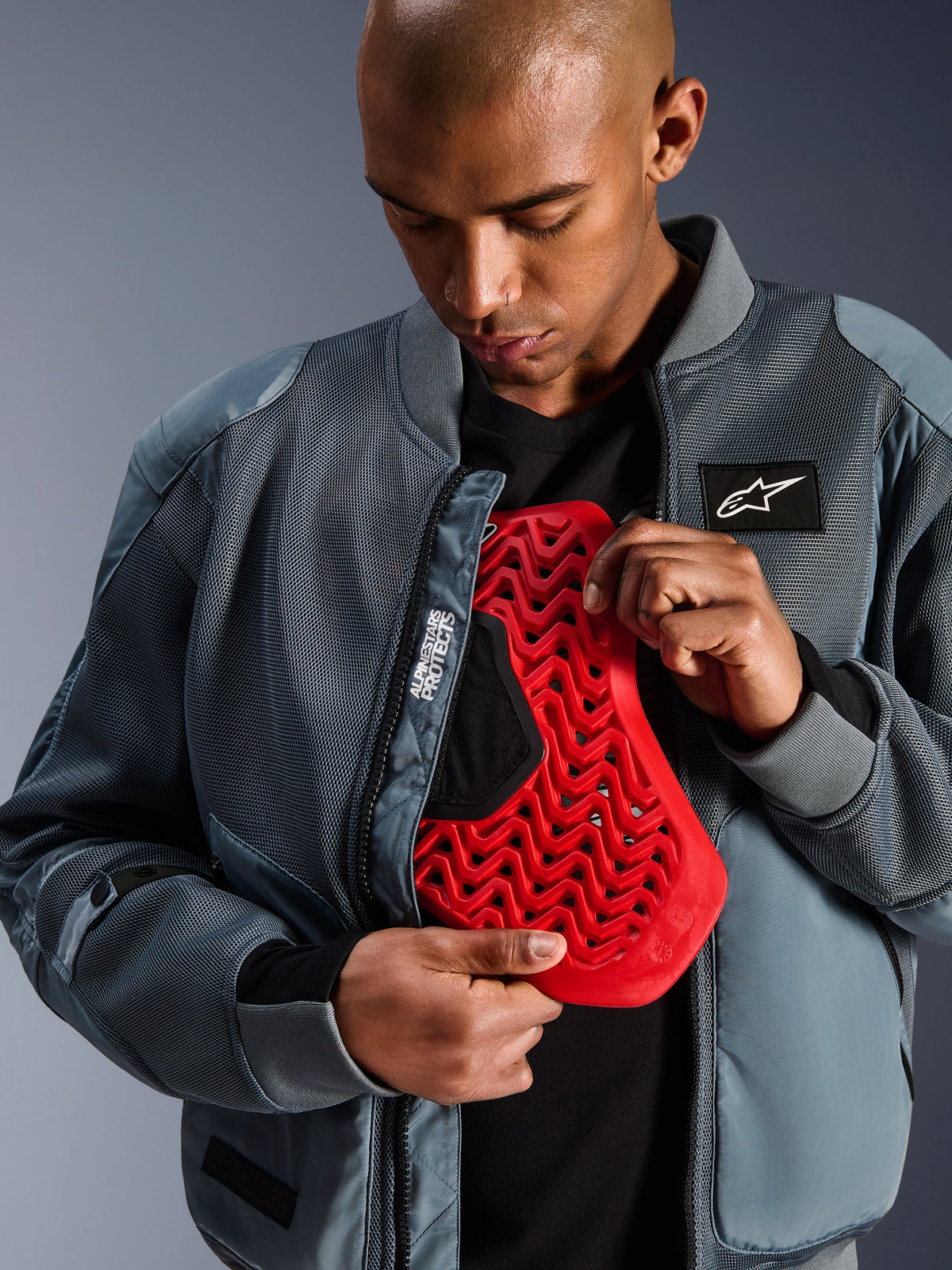 Alpinestars Flight Air Jacket in Storm Gray, urban motorcycle bomber jacket with 3D mesh panels for airflow, unzipped to reveal a red Nucleon back protector insert, featuring 'Alpinestars Protects' branding and a modern streetwear aesthetic