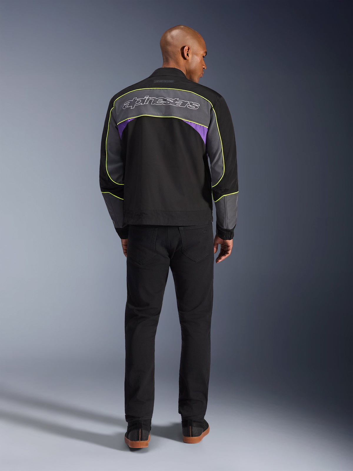 Alpinestars Unite Jacket, urban motorcycle jacket in Black Asphalt Violet, rear view showing grey back panels with purple accents and neon yellow piping, Alpinestars script logo across the shoulders, worn with black pants