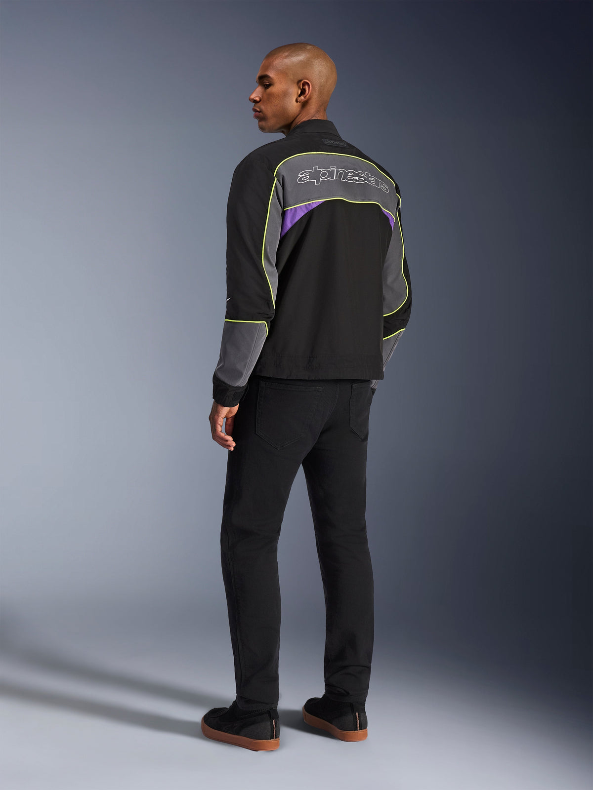 Alpinestars Unite Jacket, urban motorcycle jacket, Black Asphalt Violet, back three-quarter view on model, black softshell with grey panels and violet accents, neon yellow reflective piping, Alpinestars logo on the upper back, paired with black pants and black casual shoes