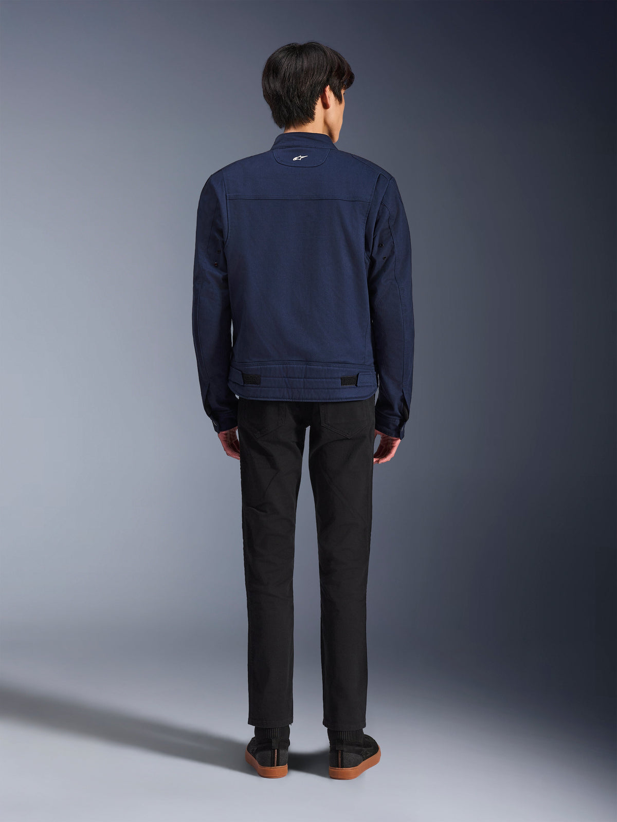 Alpinestars Bruiser Jacket in Dark Blue, back view on model, textile motorcycle jacket with a workwear-inspired design, featuring a small white logo on the back collar and adjustable waist tabs, paired with black pants