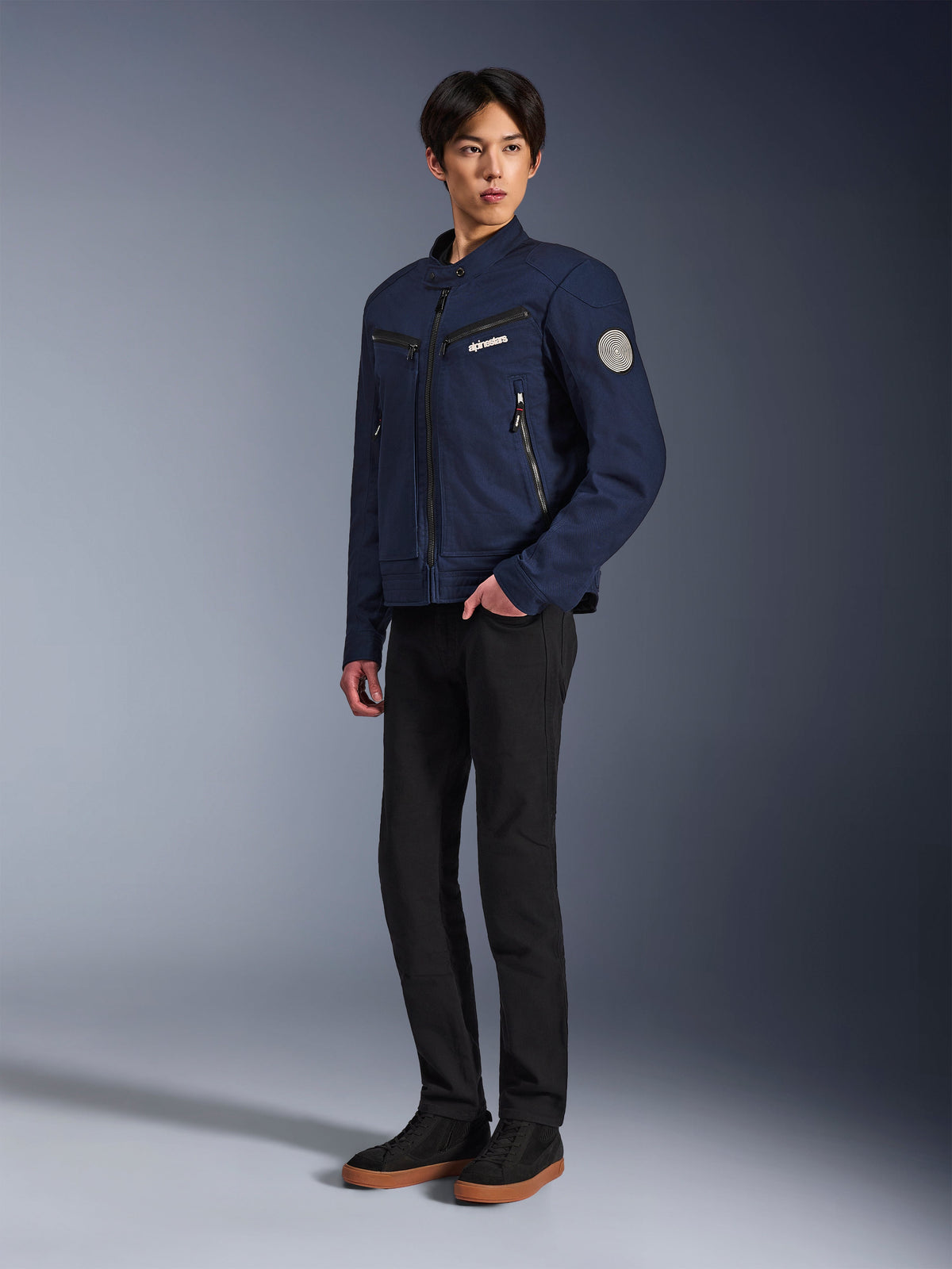 Alpinestars Bruiser Jacket in Dark Blue, urban textile motorcycle jacket with workwear-inspired design, featuring zippered chest pockets and Oscar spiral sleeve patch, worn with black pants and casual sneakers.
