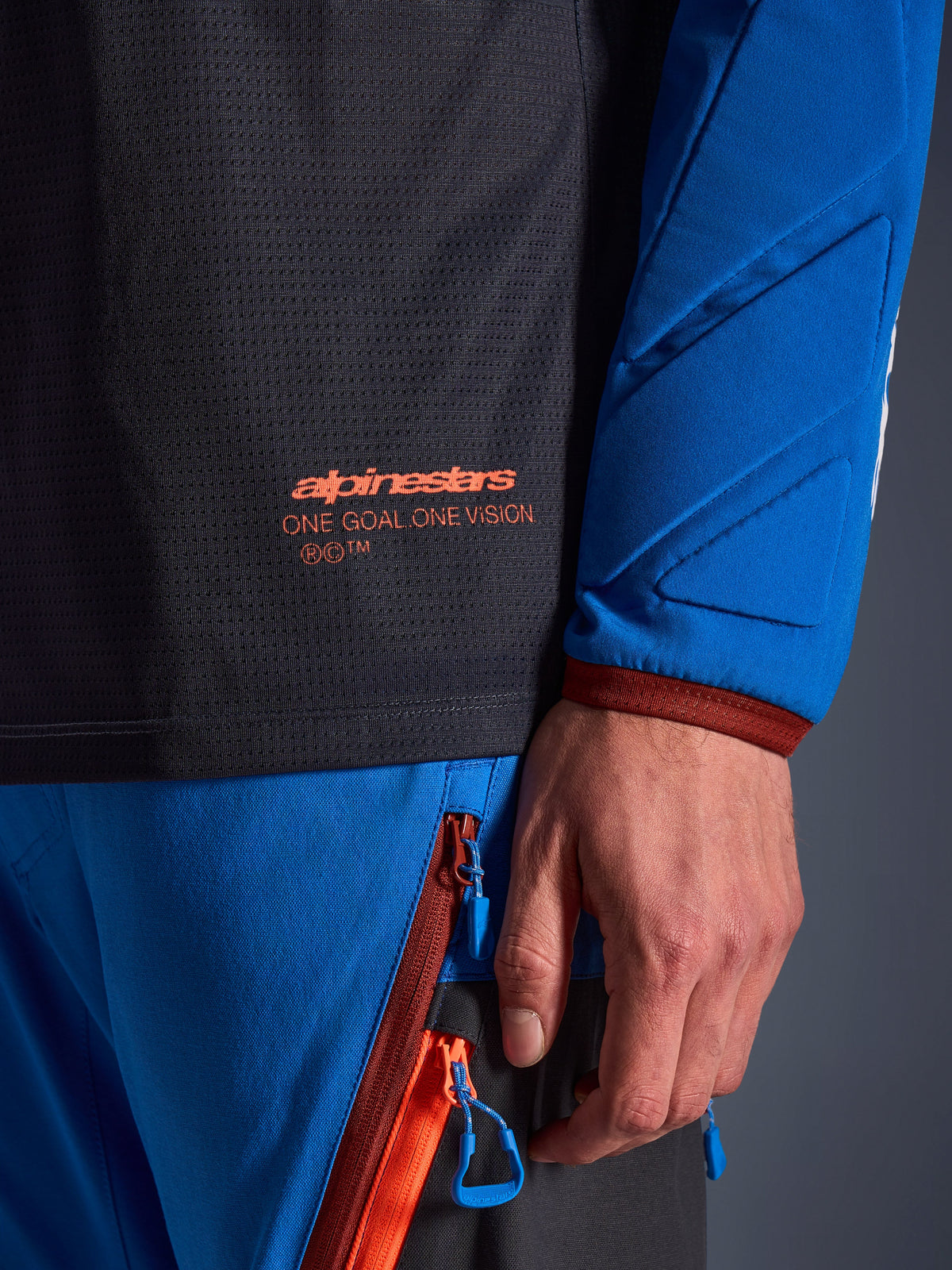 Close-up of Alpinestars Maxdura Dual Jersey in Deep Gray and Royal Blue, featuring an orange logo and 'One Goal One Vision' text on the mesh torso, padded sleeve with reinforced geometric panels, technical adventure riding gear detail