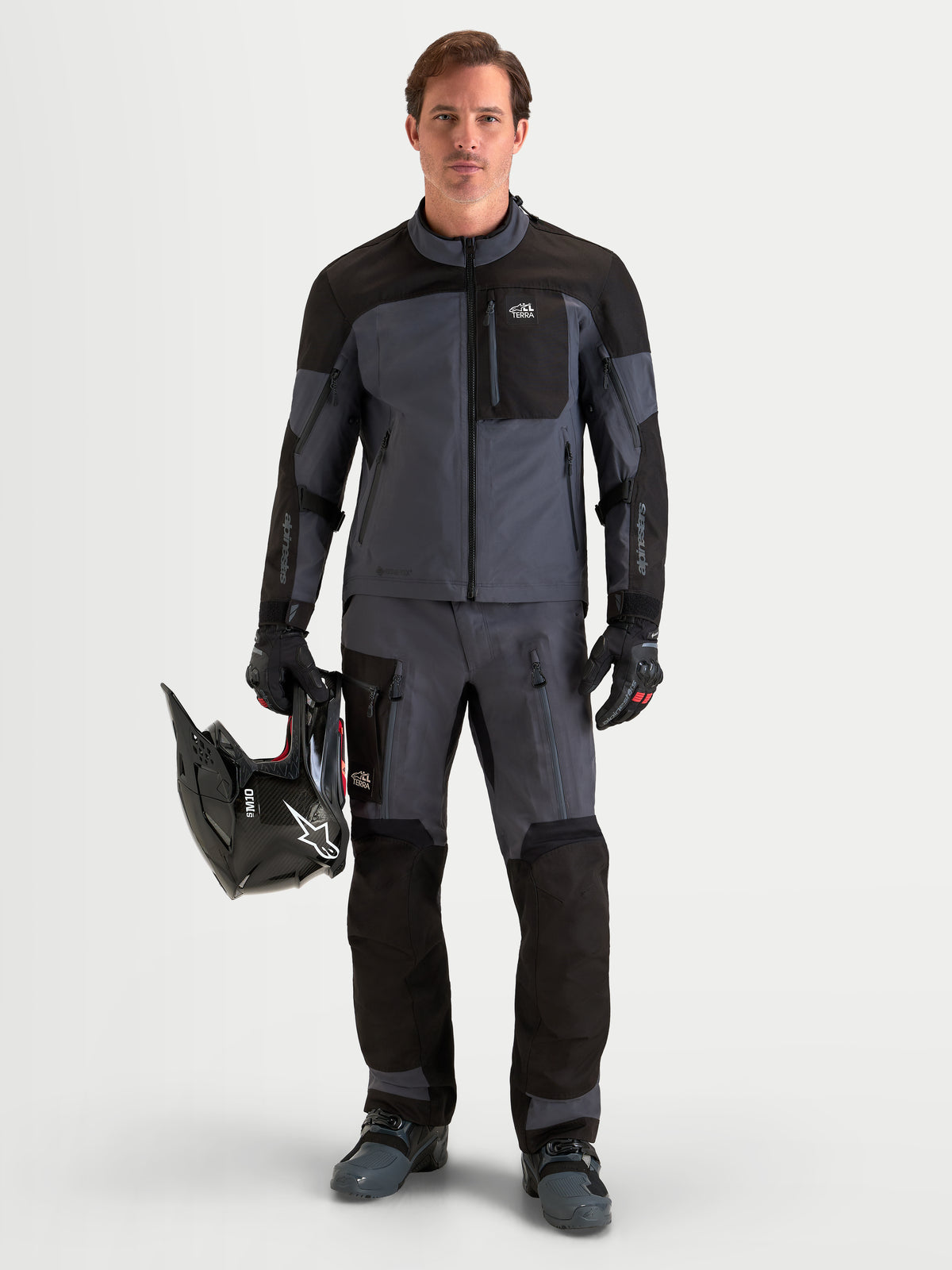 Alpinestars Arenis Gore-Tex Gear Set, men's adventure touring motorcycle jacket and pants in black and dark gray, full-length front view of a model standing while holding a helmet and wearing matching waterproof textile riding apparel.