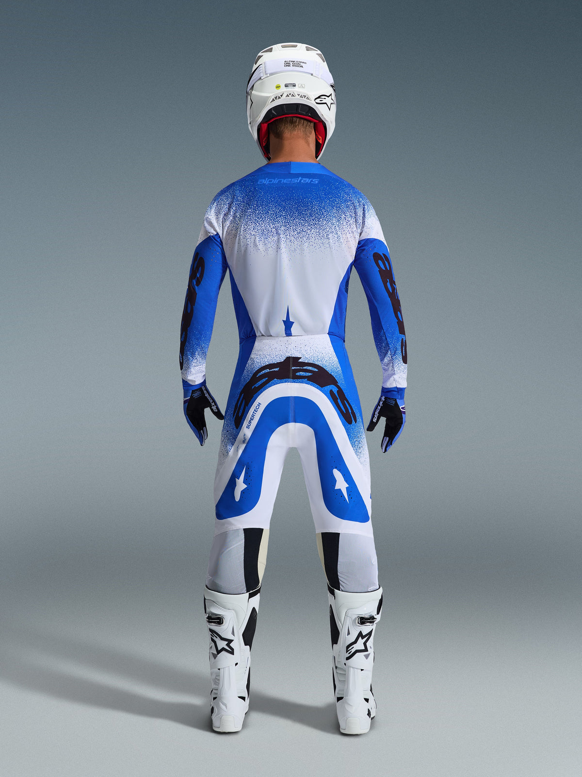 Motocross rider from behind wearing full racing gear, Alpinestars 2026 Supertech Scenz Pants in Ucla Blue and White, matching blue and white jersey with speckled pattern, white helmet, white and black boots, coordinated outfit for professional racing