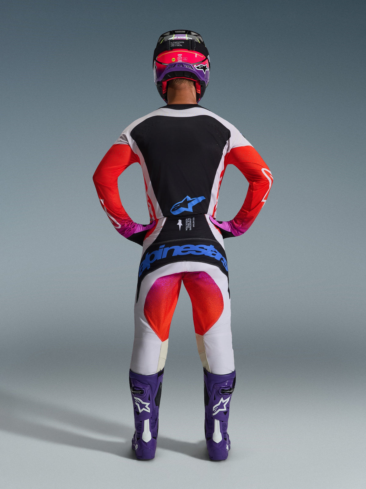 Motocross rider from behind wearing 2026 Techstar Knif Pants and matching jersey, light gray and black design with bright orange and purple accents, blue Alpinestars branding on the seat, purple boots and helmet, professional off-road racing gear set.