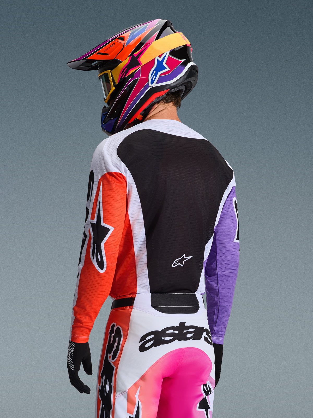Motocross rider from a rear view wearing the Alpinestars 2026 Racer Air Portl Jersey in Multicolor, featuring an orange right sleeve, purple left sleeve, and a large black mesh back panel for ventilation, paired with matching motocross pants and a vibrant multi-colored helmet, professional off-road racing equipment.