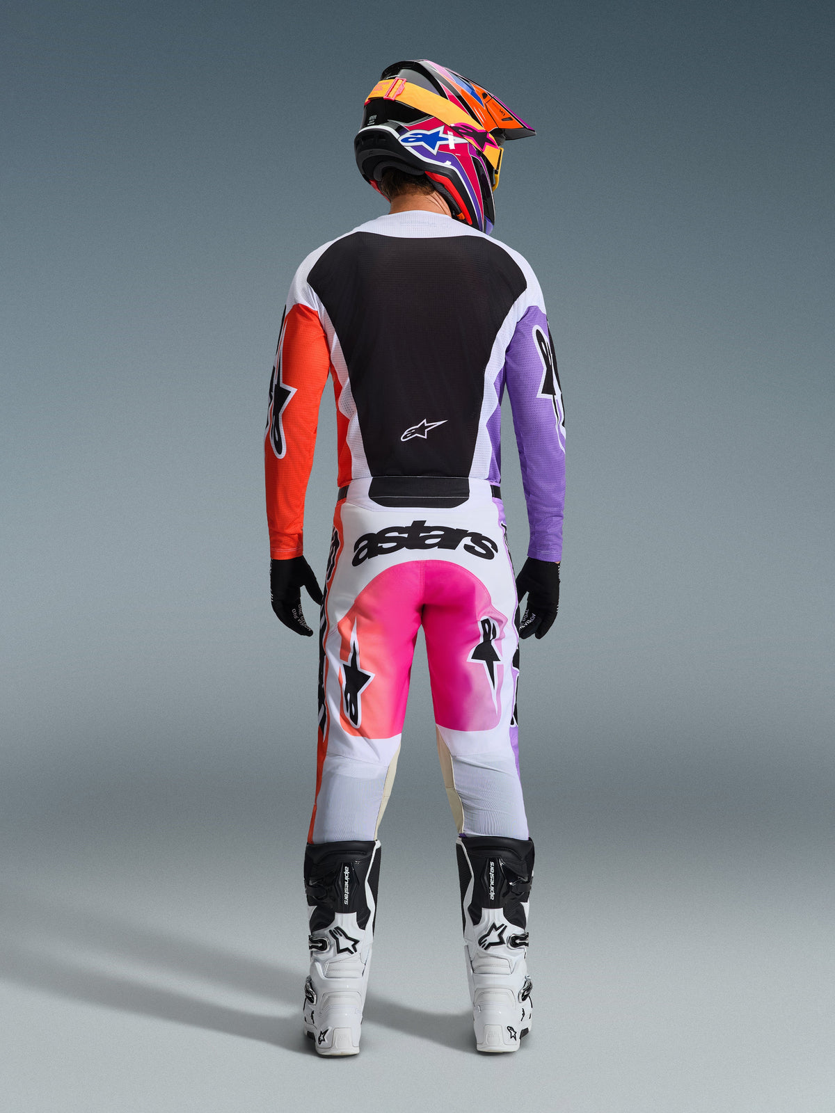 Motocross rider wearing complete racing gear from the rear, Alpinestars 2026 Racer Air Portl Jersey with white, black, orange, and purple panels, matching white pants with pink accents and 'astars' branding, white and black boots, and a vibrant multicolor helmet, coordinated outfit for motocross racing