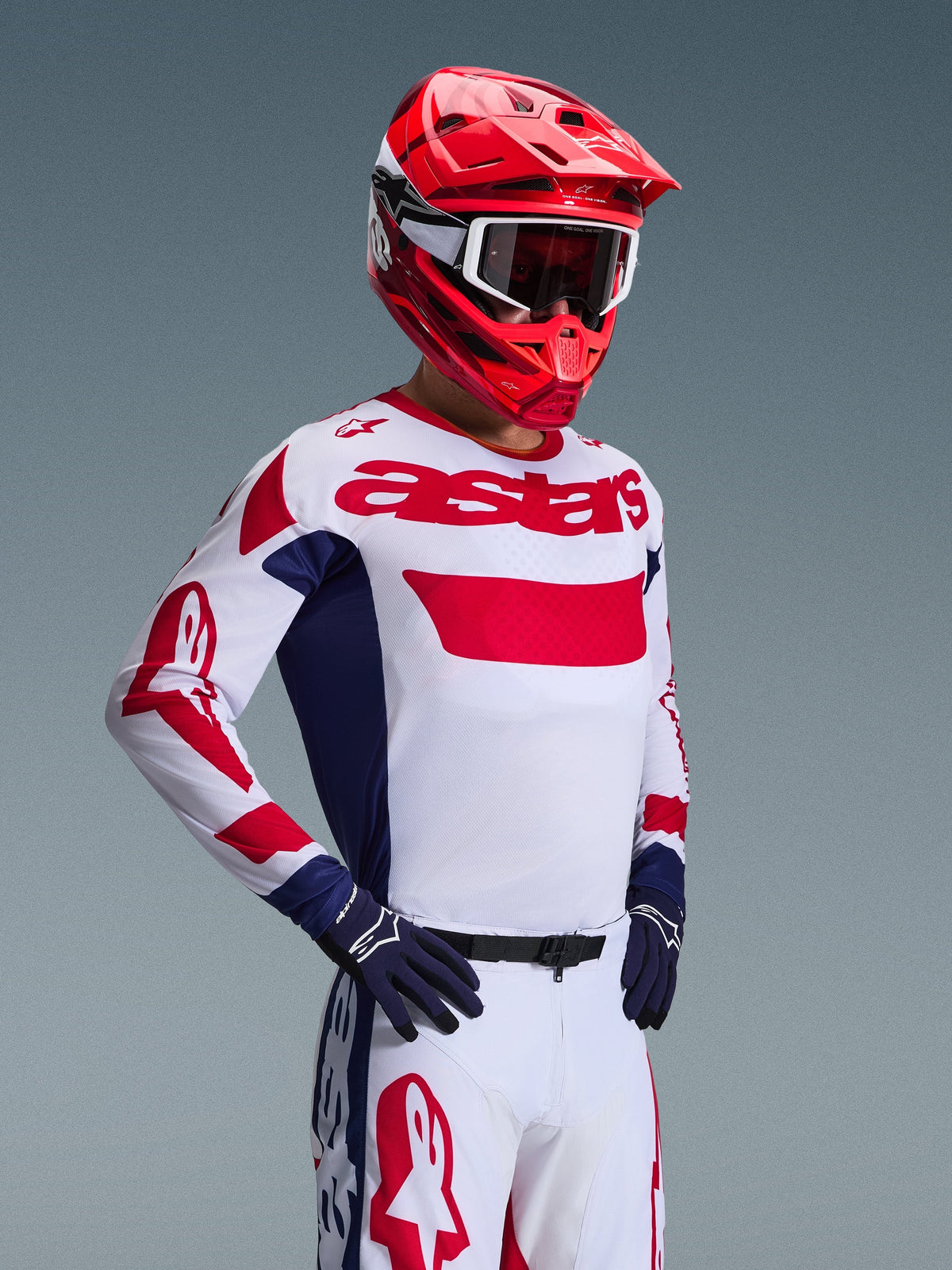 Motocross rider wearing the Alpinestars 2026 Racer Riway Jersey in White Blue Red, featuring bold red 'astars' branding on the chest and blue side panels, paired with a matching red helmet and white racing pants for a complete off-road performance look.