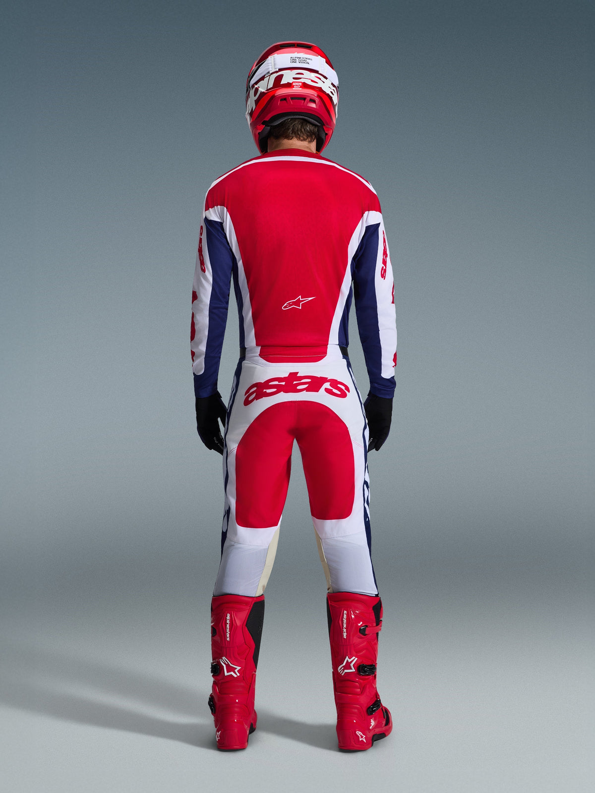 Motocross rider wearing complete racing gear from behind, Alpinestars 2026 Racer Riway Jersey in White Blue Red, matching white and red pants with bold 'astars' branding, red boots, and red and white helmet, coordinated off-road racing outfit.