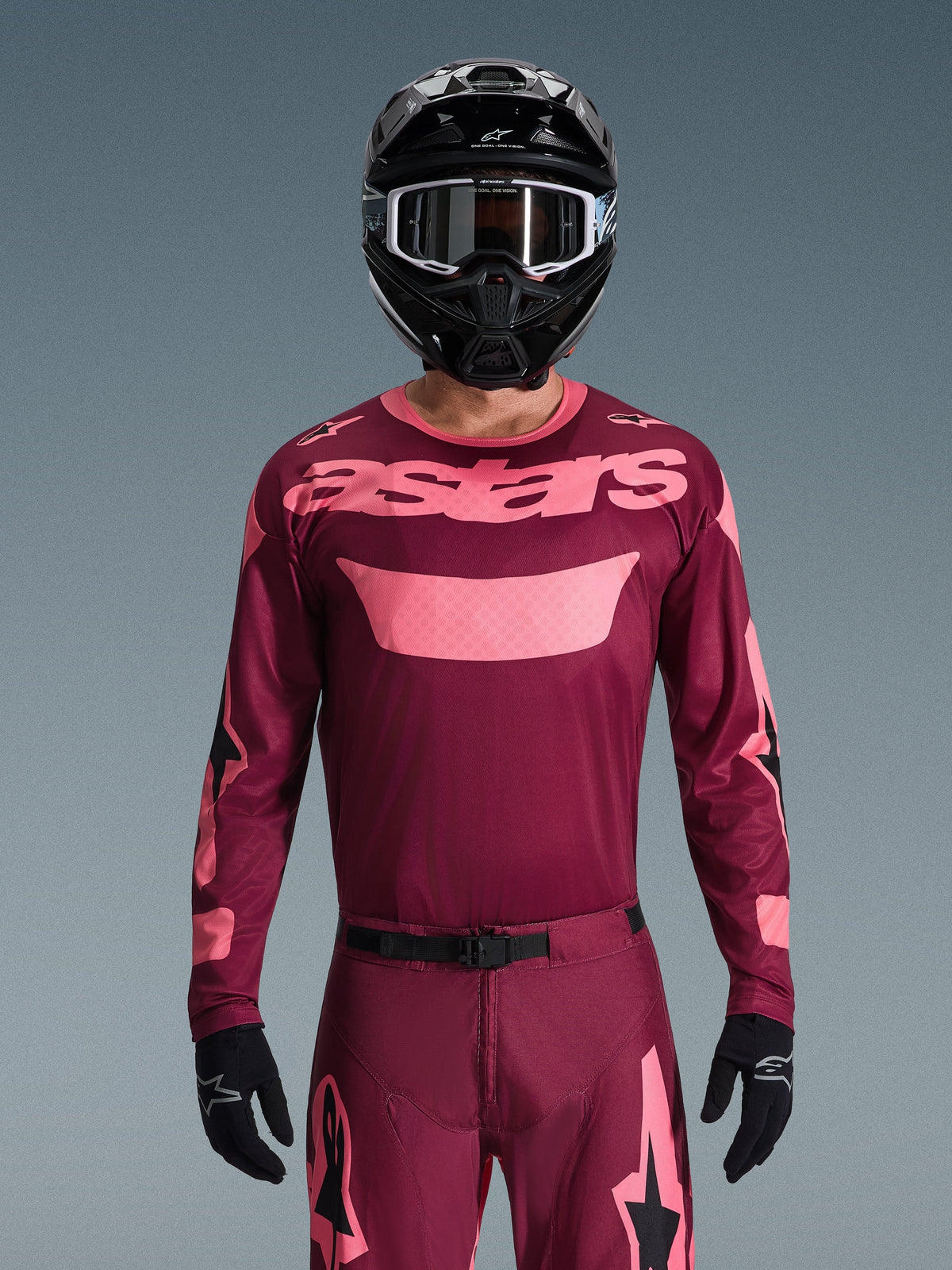 Motocross rider wearing the Alpinestars 2026 Racer Riway Jersey in Dark Red Coral, featuring a maroon body with coral pink 'astars' chest branding, paired with matching motocross pants, black helmet, and black gloves, professional gear set for off-road racing.