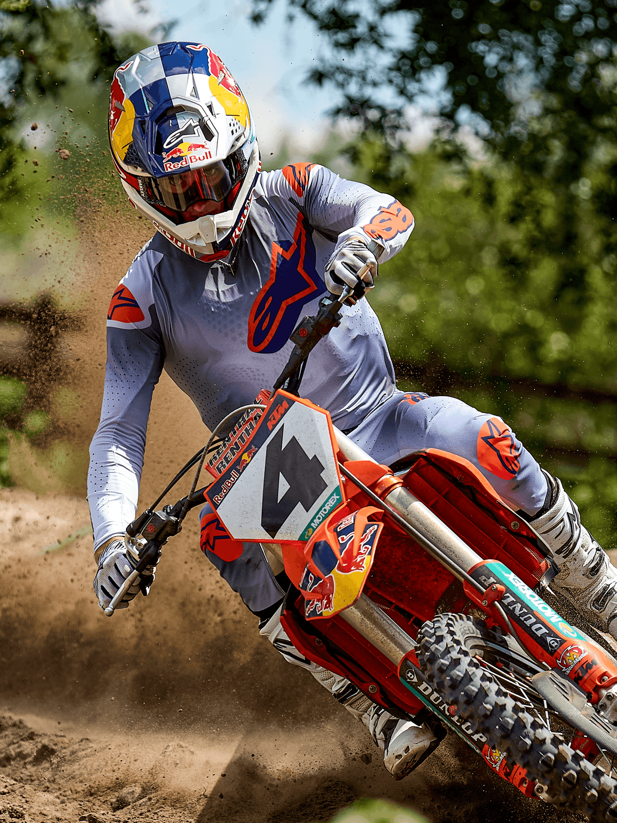 Motocross rider in action on a dirt track wearing the Alpinestars 2025 Supertech Lipan Jersey in Gray Orange Fluo, matching gray pants with orange accents, white boots, and a Red Bull branded helmet, cornering on an orange dirt bike with dirt spray, professional off-road racing gear