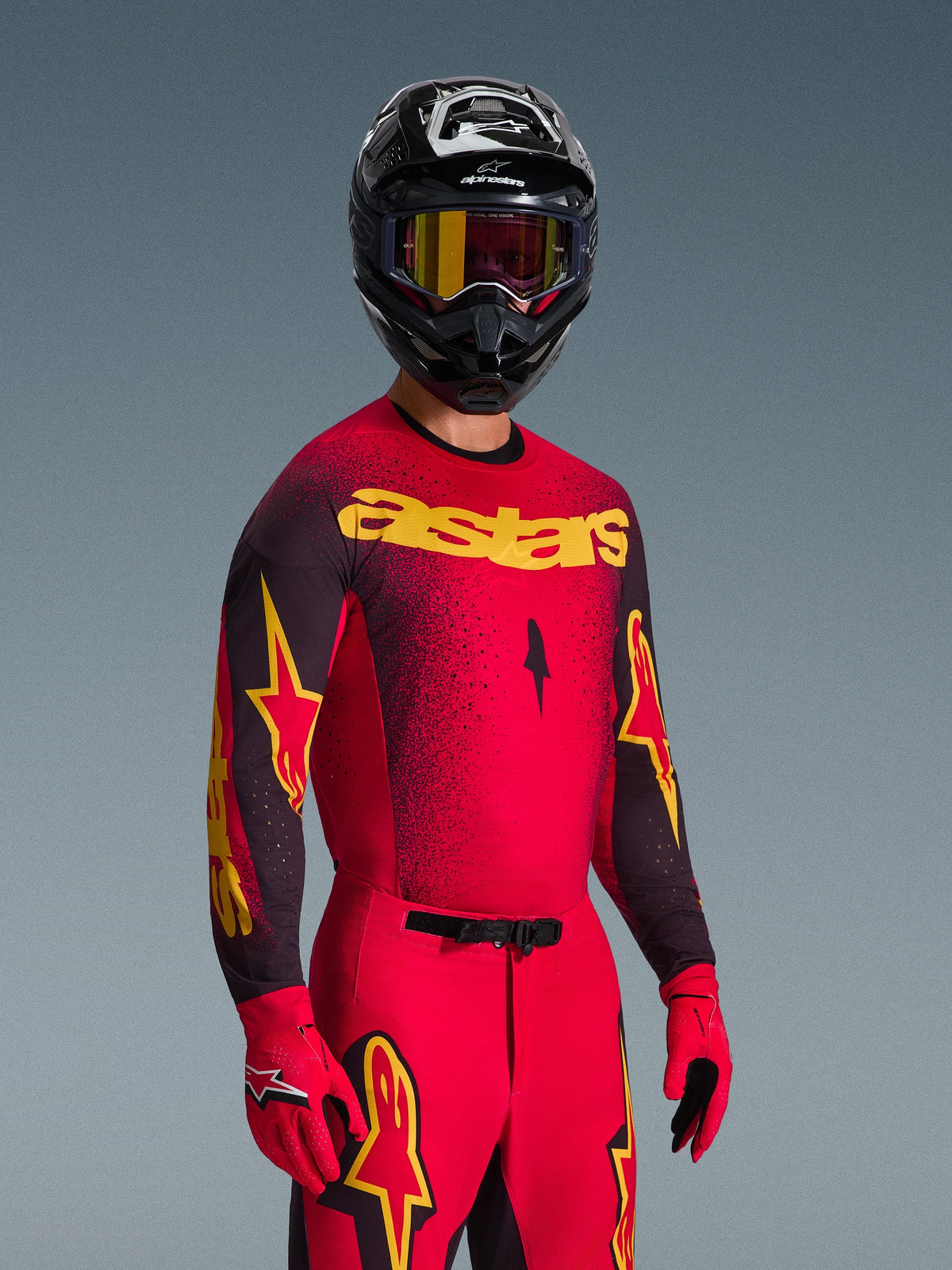 Motocross rider wearing Alpinestars 2026 Supertech Scenz Jersey in red and black with yellow 'astars' branding, matching red pants and gloves, black helmet with goggles, professional racing gear set for off-road motocross