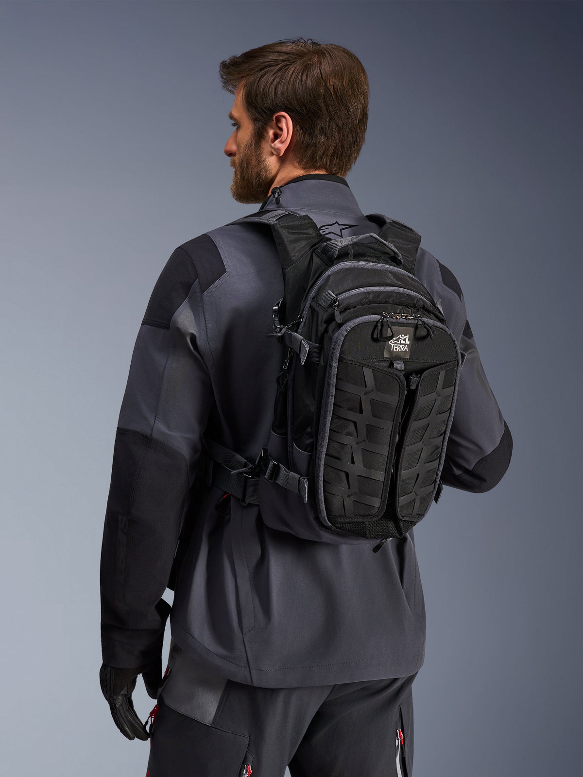 Alpinestars AT-15 Back Pack, Adventure Motorcycle Backpack, Black and Deep Gray, worn by a rider in a gray jacket, featuring a technical design with MOLLE webbing and 'TERRA' branding, ergonomic off-road gear