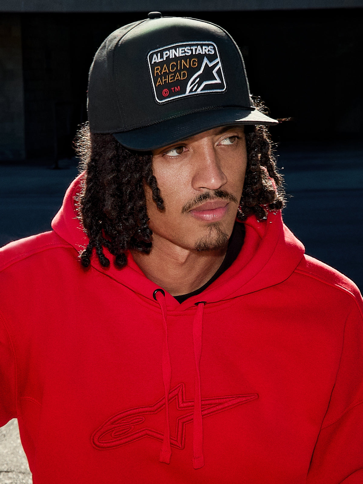 Man wearing Alpinestars Intuitive Snapback Hat in Black, featuring a structured crown and 'Racing Ahead' logo embroidery patch, paired with a bright red hoodie, casual lifestyle streetwear