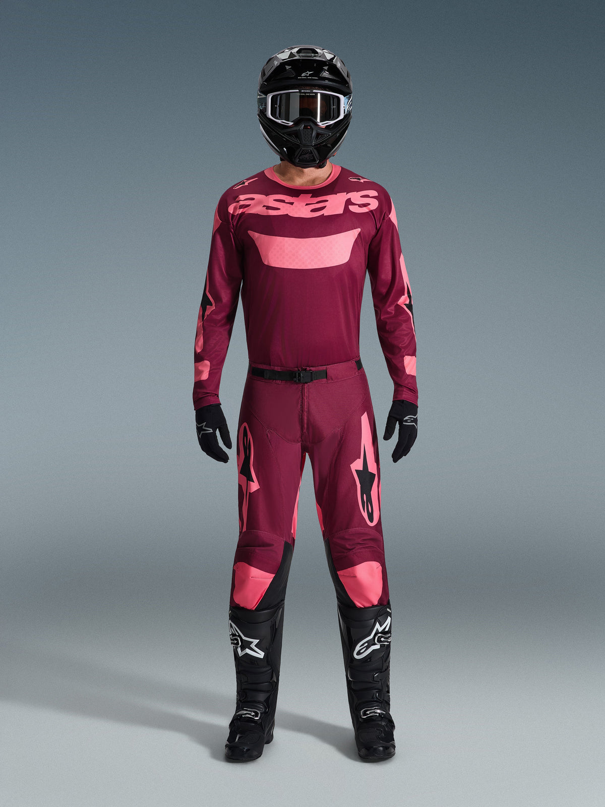 Motocross rider wearing the Alpinestars 2026 Racer Riway Gear Set, maroon jersey with pink 'astars' branding, matching maroon pants with pink knee and leg accents, black helmet, black gloves, and black boots, complete performance outfit for off-road racing