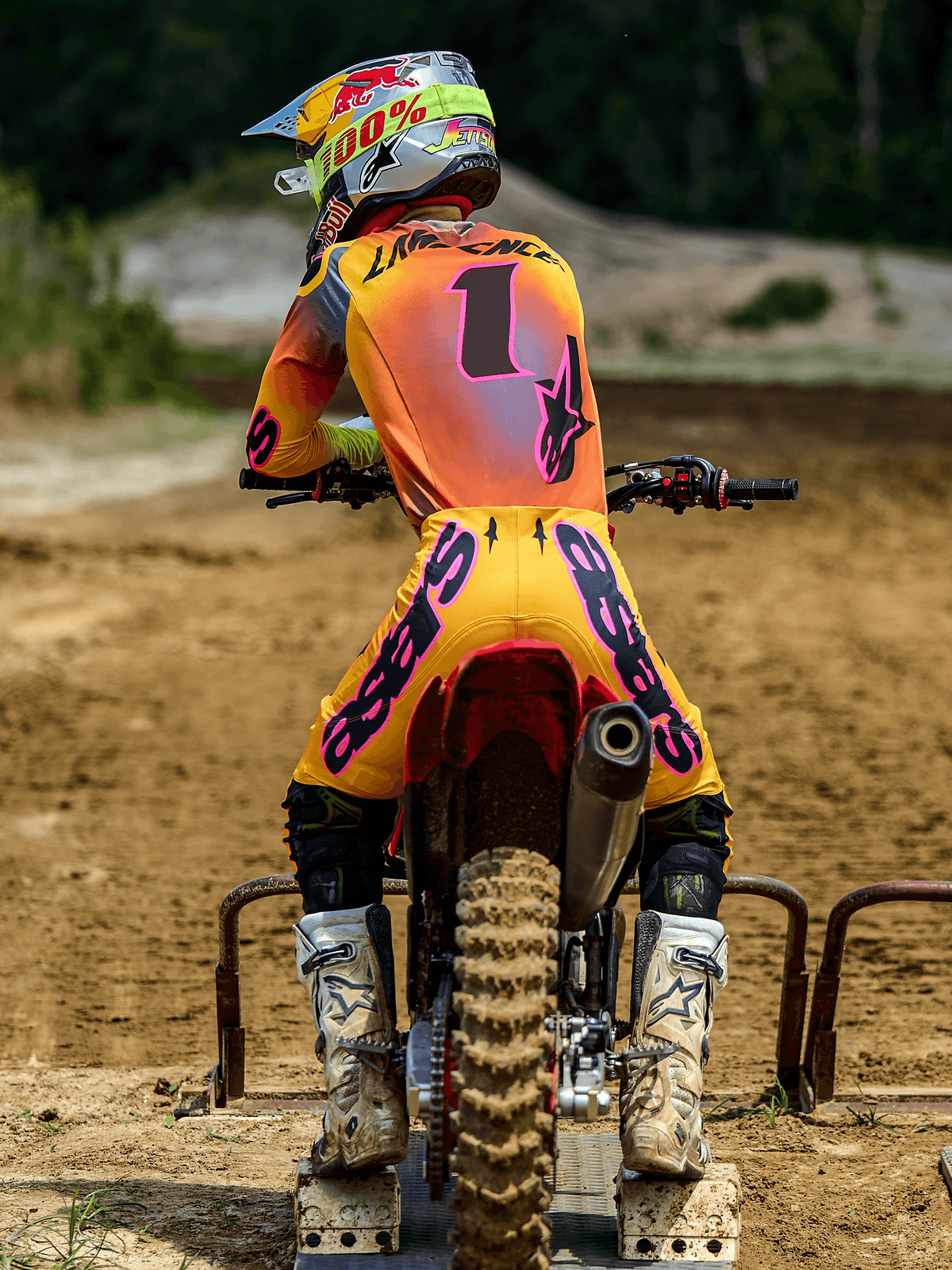 Motocross rider viewed from behind at a starting gate, wearing Alpinestars 2025 Supertech Lipan Pants in Yellow and Pink, matching jersey with pink accents and number 1, white Alpinestars boots, and a racing helmet, coordinated professional off-road racing gear