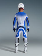 Motocross rider from behind wearing full racing gear, Alpinestars 2026 Supertech Scenz Pants in Ucla Blue and White, matching blue and white jersey with speckled pattern, white helmet, white and black boots, coordinated outfit for professional racing