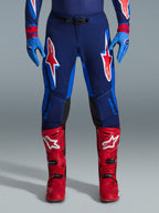 Alpinestars 2026 Supertech Vista Pants, Motocross Pants, Dark Navy, Hot Coral, and Cobalt, front view of rider wearing performance gear with matching blue gloves and red motocross boots, featuring prominent red and white Alpinestars logos on thighs.