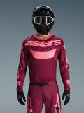 Motocross rider wearing the Alpinestars 2026 Racer Riway Jersey in Dark Red Coral, featuring a maroon body with coral pink 'astars' chest branding, paired with matching motocross pants, black helmet, and black gloves, professional gear set for off-road racing.