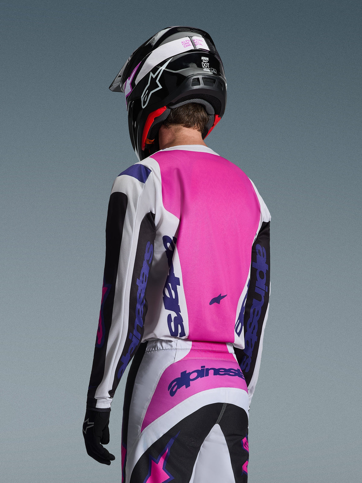 Motocross rider seen from the back wearing the Alpinestars 2026 Fluid Grid Jersey in Black, White, and Purple, matching pants with bold branding, black helmet with pink accents, and black gloves, complete coordinated gear for off-road performance.