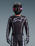 Alpinestars 2026 Fluid Apex Jersey, Motocross Jersey, Black and Gray, featuring a large white Astars logo on the chest and gray graphic accents on the sleeves, worn by a rider with pink goggles and a black helmet, coordinated off-road racing gear