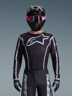 Alpinestars 2026 Fluid Apex Jersey, Motocross Jersey, Black and Gray, featuring a large white Astars logo on the chest and gray graphic accents on the sleeves, worn by a rider with pink goggles and a black helmet, coordinated off-road racing gear