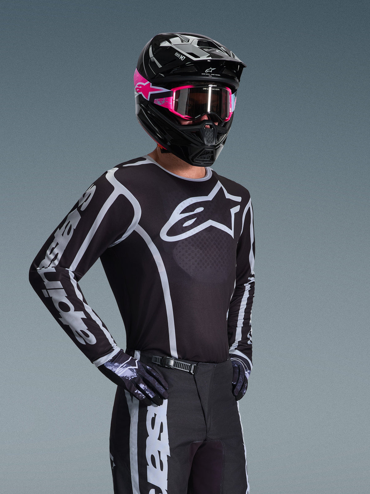 Motocross rider wearing the Alpinestars 2026 Fluid Apex Jersey, Black and Gray with large white chest logo and geometric graphics on sleeves, paired with black pants, black gloves, and a black helmet with pink accents and pink goggles
