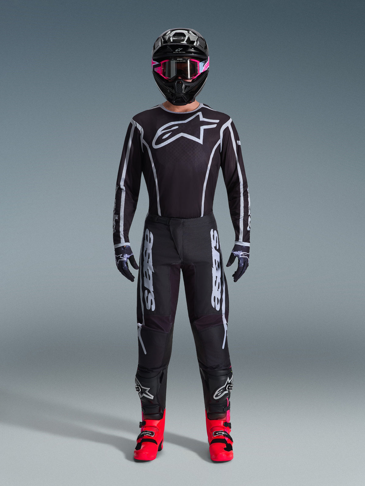 Motocross rider wearing the Alpinestars 2026 Fluid Apex Jersey, Black and Gray, large white Astars logo on the chest, white geometric lines, matching black pants, black helmet with pink goggles, and bright pink boots, full motocross racing outfit