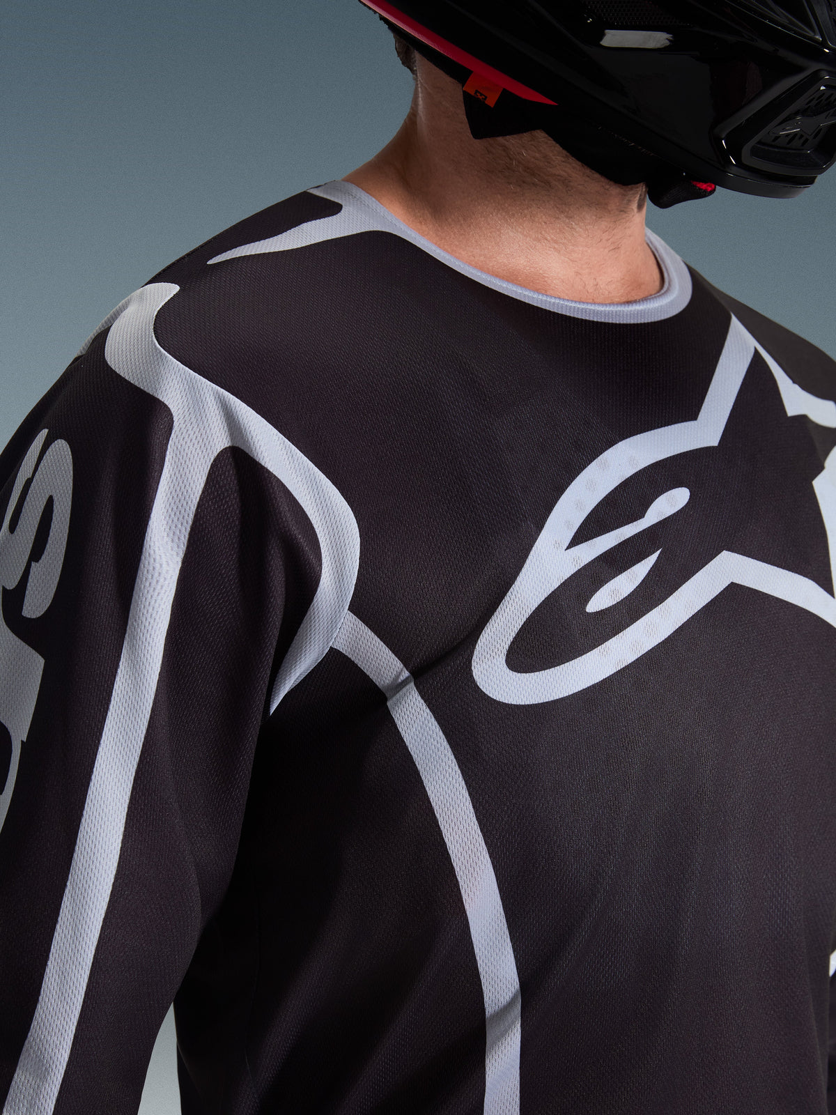 Close-up of a rider wearing the Alpinestars 2026 Fluid Apex Jersey in Black and Gray, showcasing the large white Astars logo on the chest and technical mesh fabric construction, paired with a black motocross helmet.