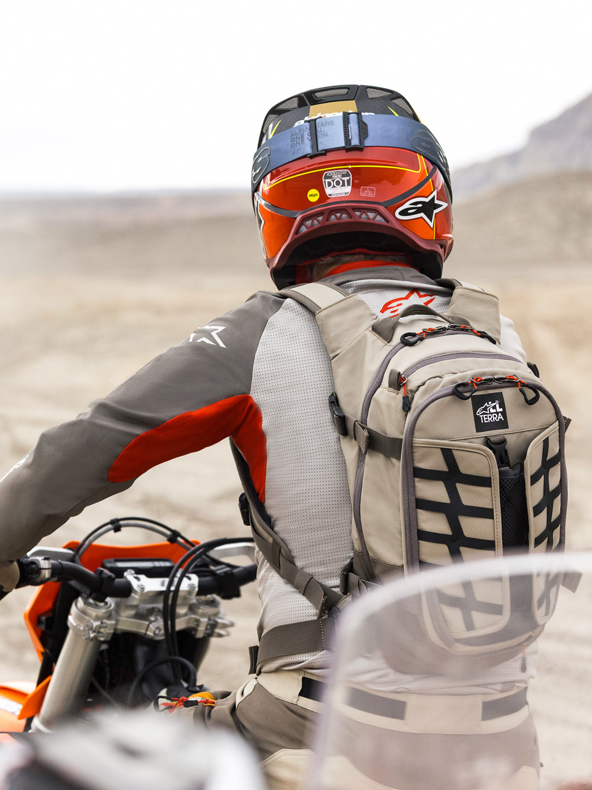 Alpinestars AT-15 Back Pack in Aluminum Walnut, worn by an adventure motorcycle rider in a desert landscape. Features rugged beige textile construction with black webbing, orange zipper accents, and 'All Terra' branding. Paired with a red and orange helmet and grey riding gear for off-road touring.