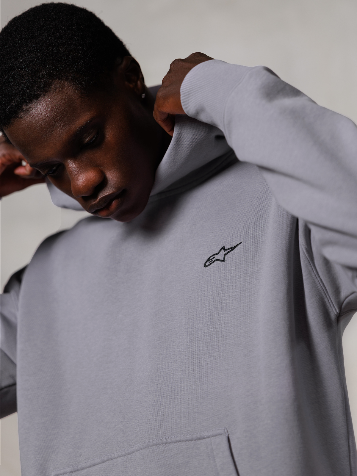 Alpinestars Essentials Ageless Hoodie, Silver, close-up of a man adjusting the hood of the grey sweatshirt, small black Alpinestars logo on the chest, premium motorsport-inspired casual wear