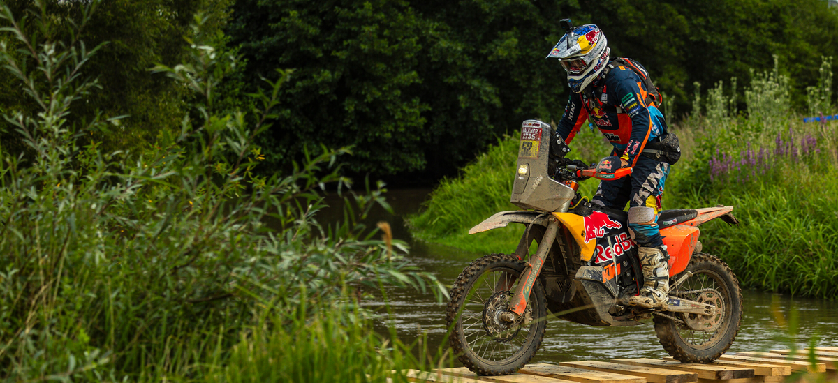 Matthias Walkner riding a dirt bike on a wooden platform over water with greenery in the background