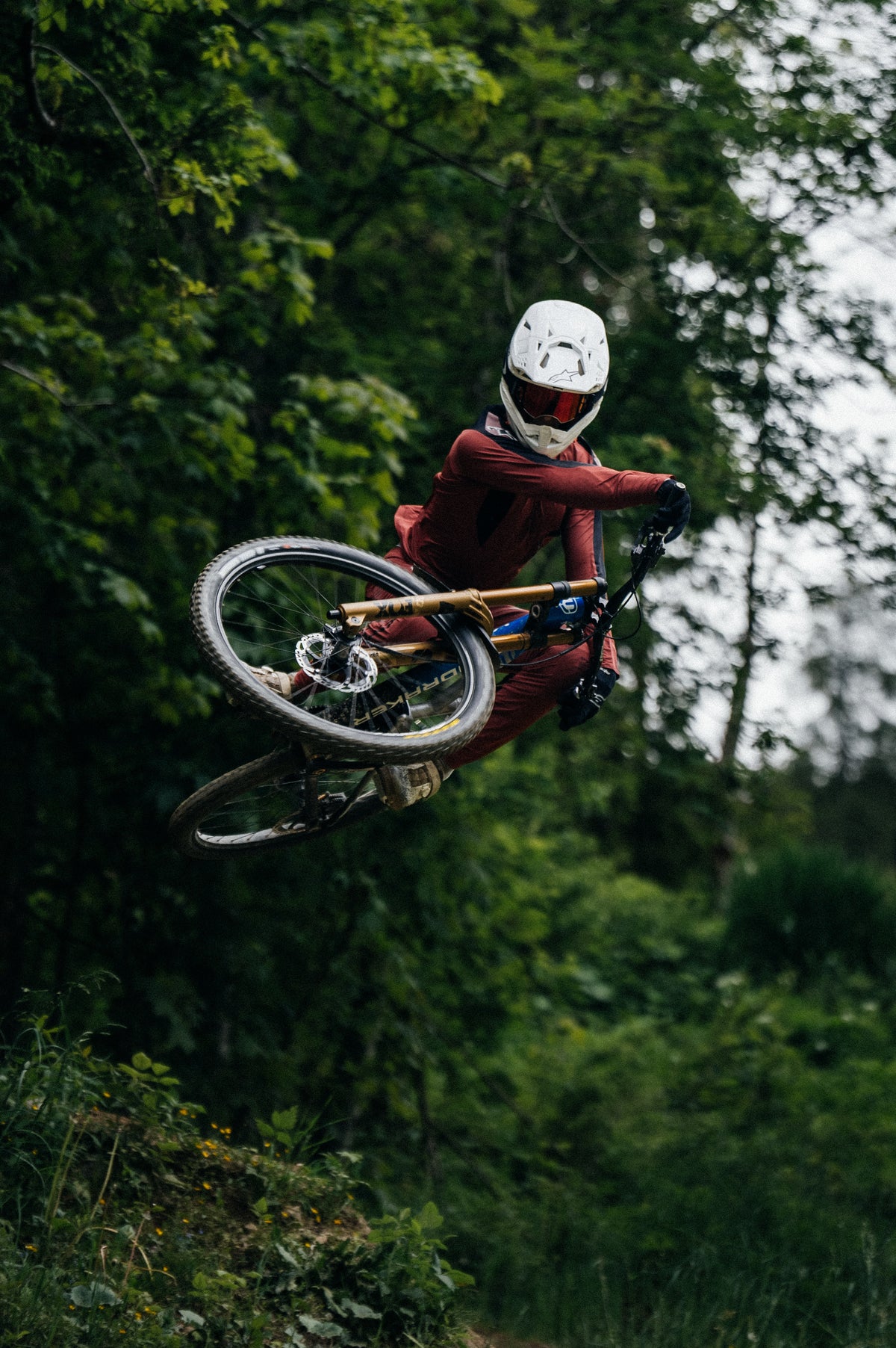 Burgundy Alpinestars A-Supra Race Jersey and Pants on a mountain biker mid-air during an off-road forest ride.